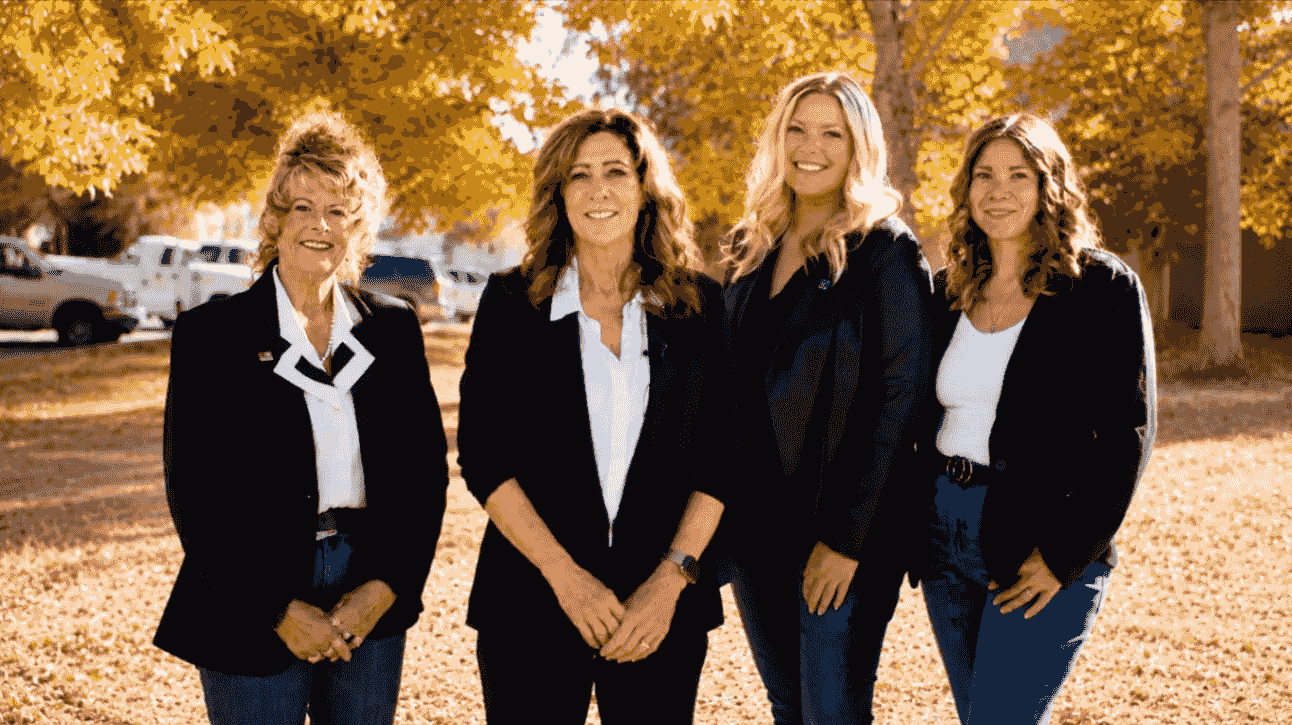 Four women in black blazers standing together outdoors among autumn trees